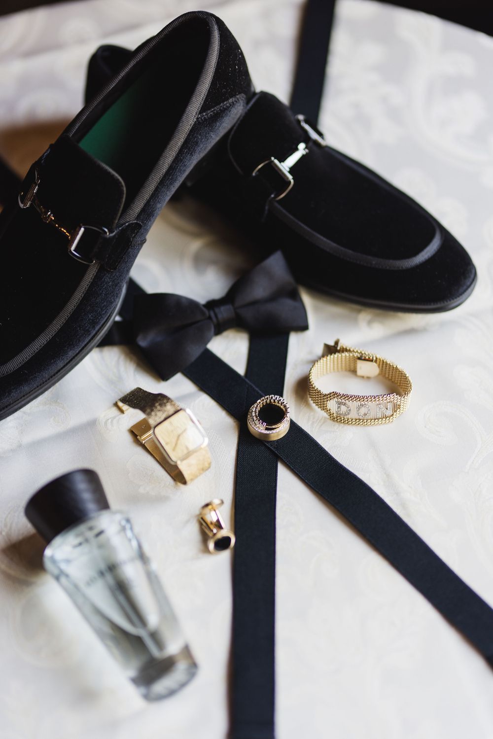 Black velvet loafers, bow tie, suspenders, gold watch, bracelet, ring, cufflinks, and cologne arranged as groom accessories.