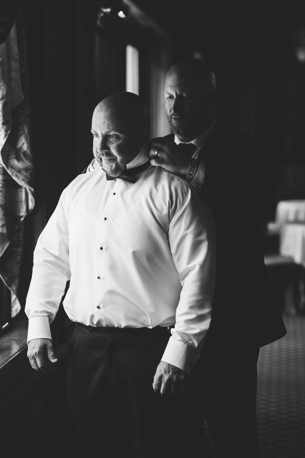 Black and white photo of a bald groom having his bow tie adjusted by his best man before the wedding.