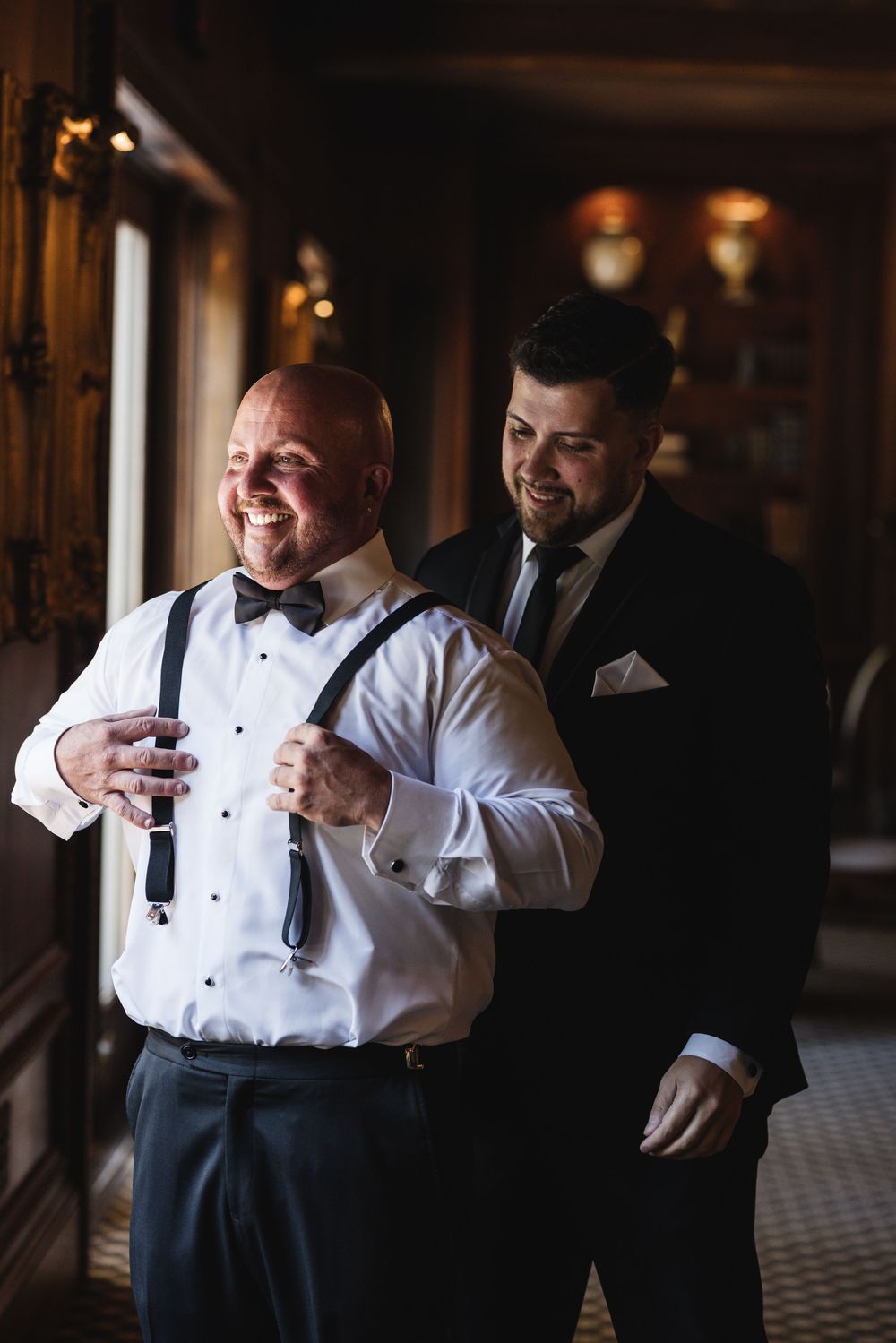 Two men in formal attire smile as they prepare for a wedding, one adjusting the other's suspenders indoors.