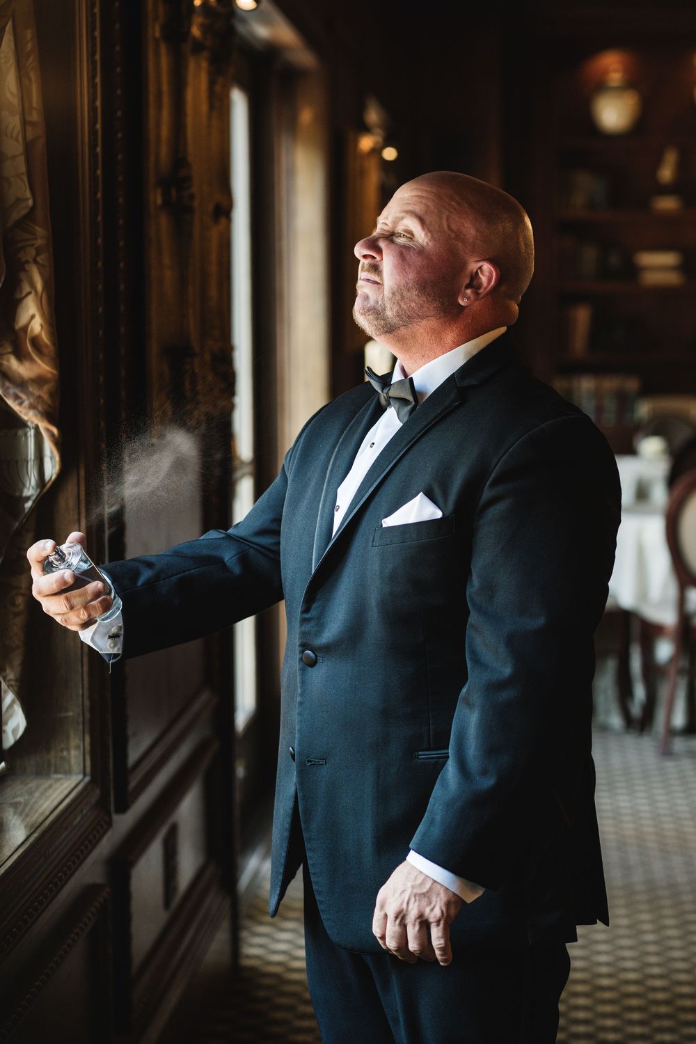 Bald groom in black tuxedo with bow tie holding spray bottle, looking up in elegant wood-paneled room.
