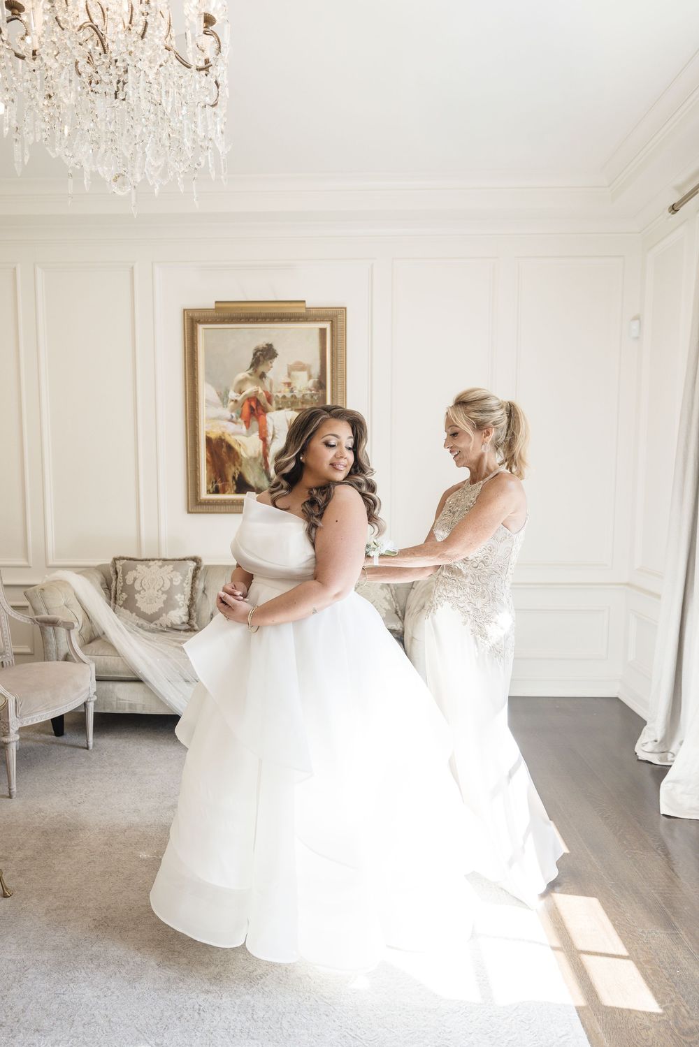 A bride in a white ballgown gets help from her bridesmaid in an elegant white room with crystal chandelier.