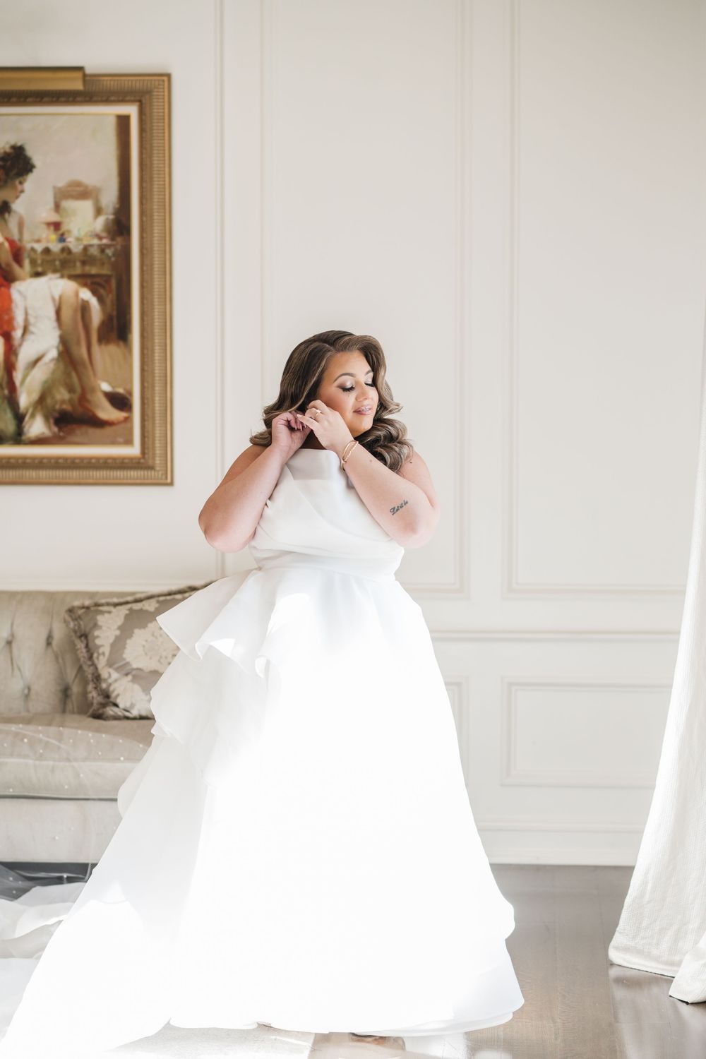 Bride in white ball gown adjusting earring in elegant white room with framed artwork.