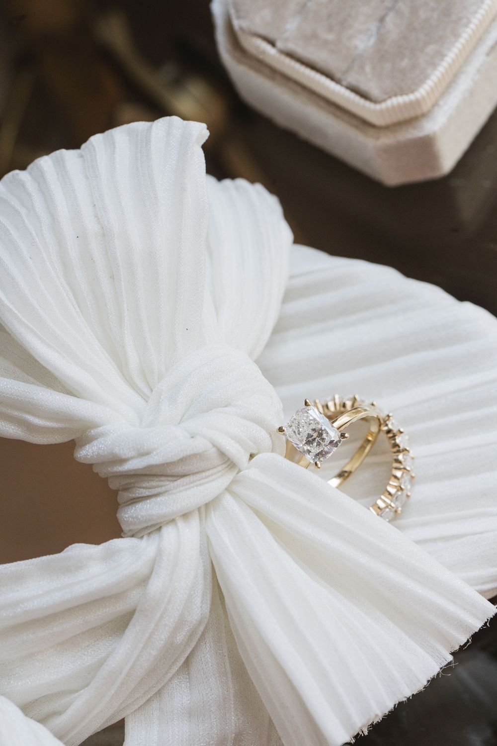 Gold diamond engagement ring resting on a white pleated ribbon bow with a velvet ring box in the background.