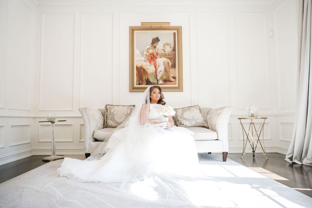 Bride in white ball gown and veil sits on elegant sofa beneath classic painting in bright white room.