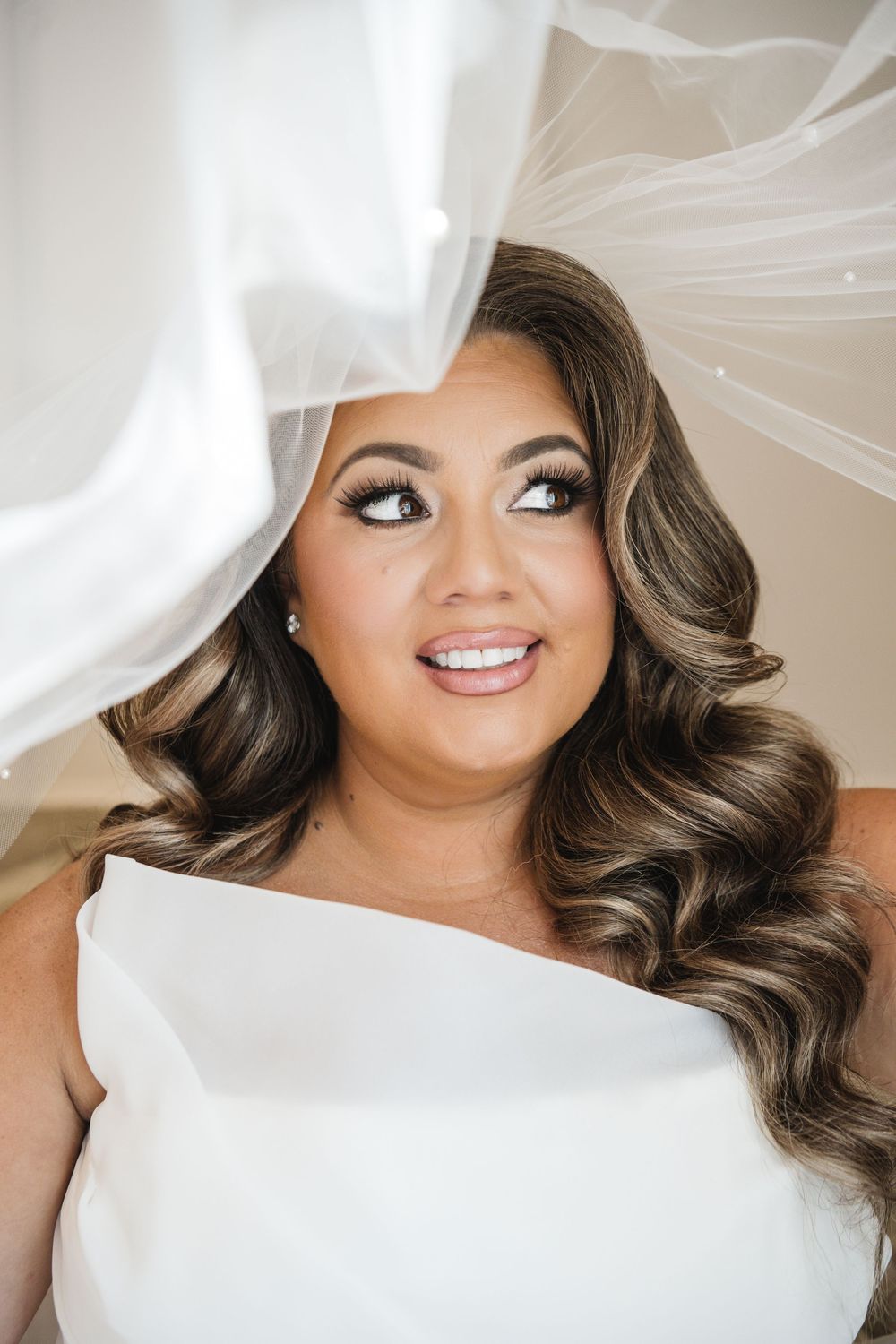 Beautiful bride with glamorous curled hair and dramatic makeup smiles under a flowing white wedding veil.