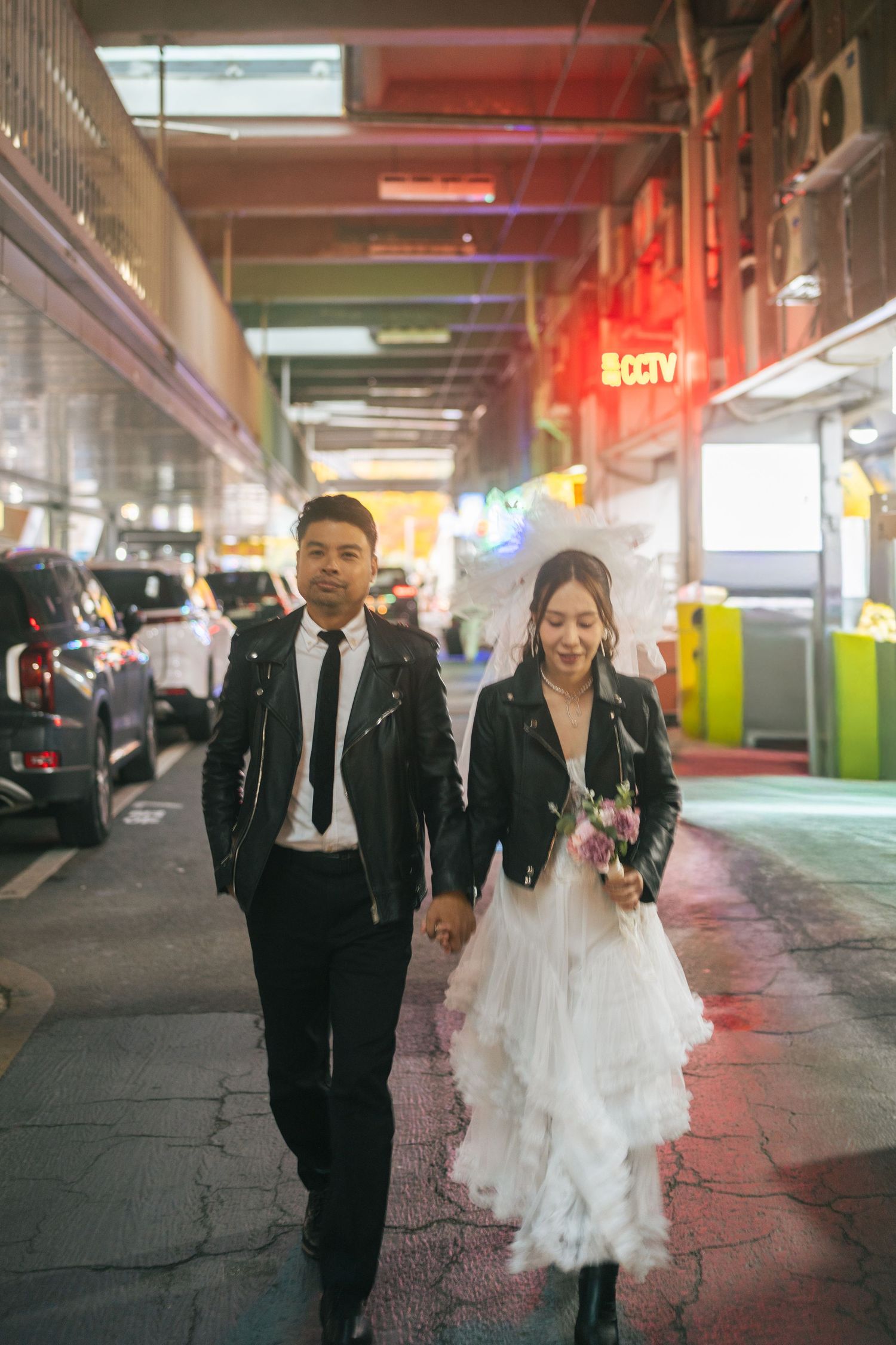 A couple in formal attire walks through a colorful parking garage with neon lighting.