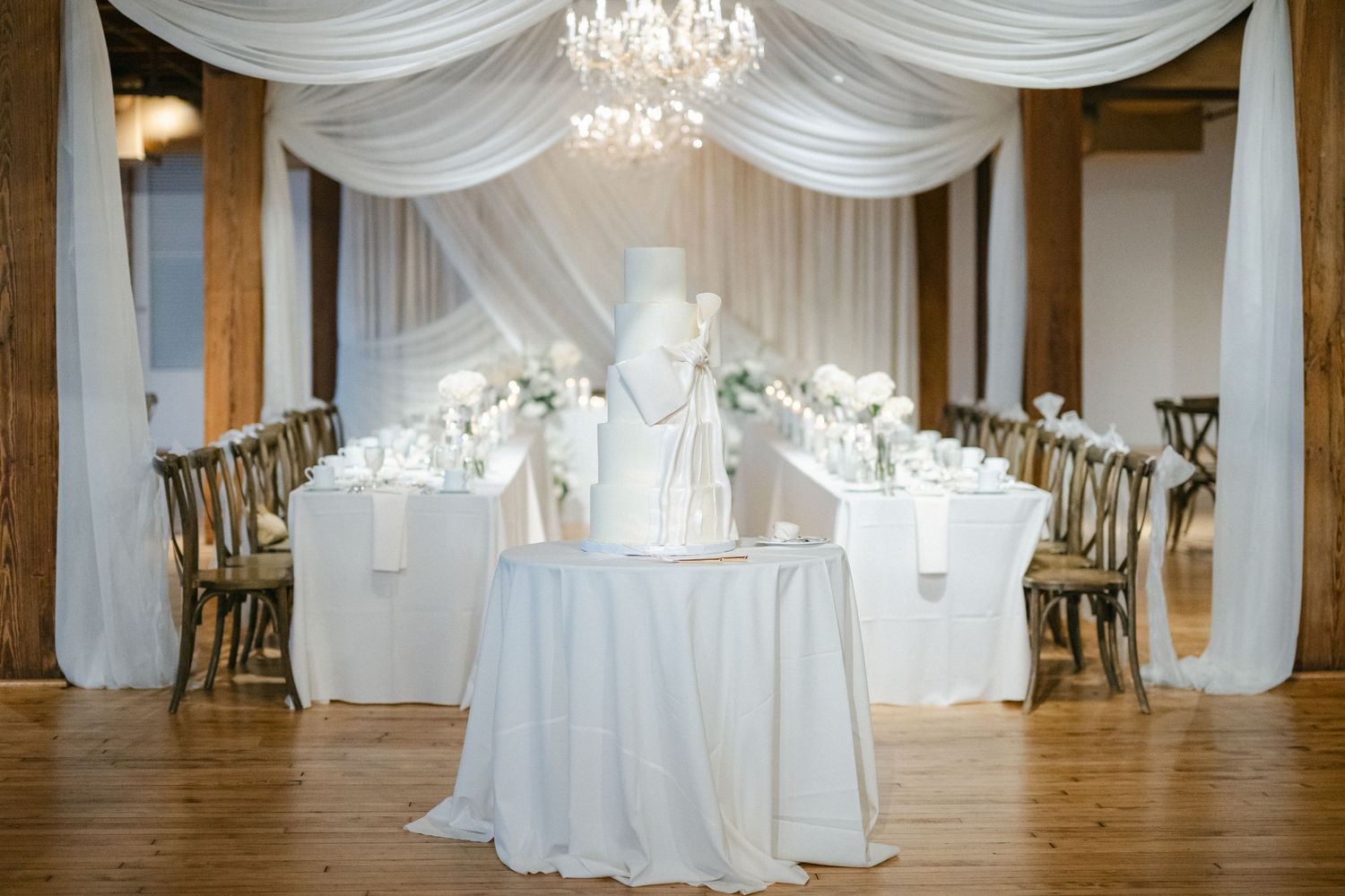 Elegant wedding reception venue with white drapes, chandeliers, rustic wood beams and cross-back chairs at tables.