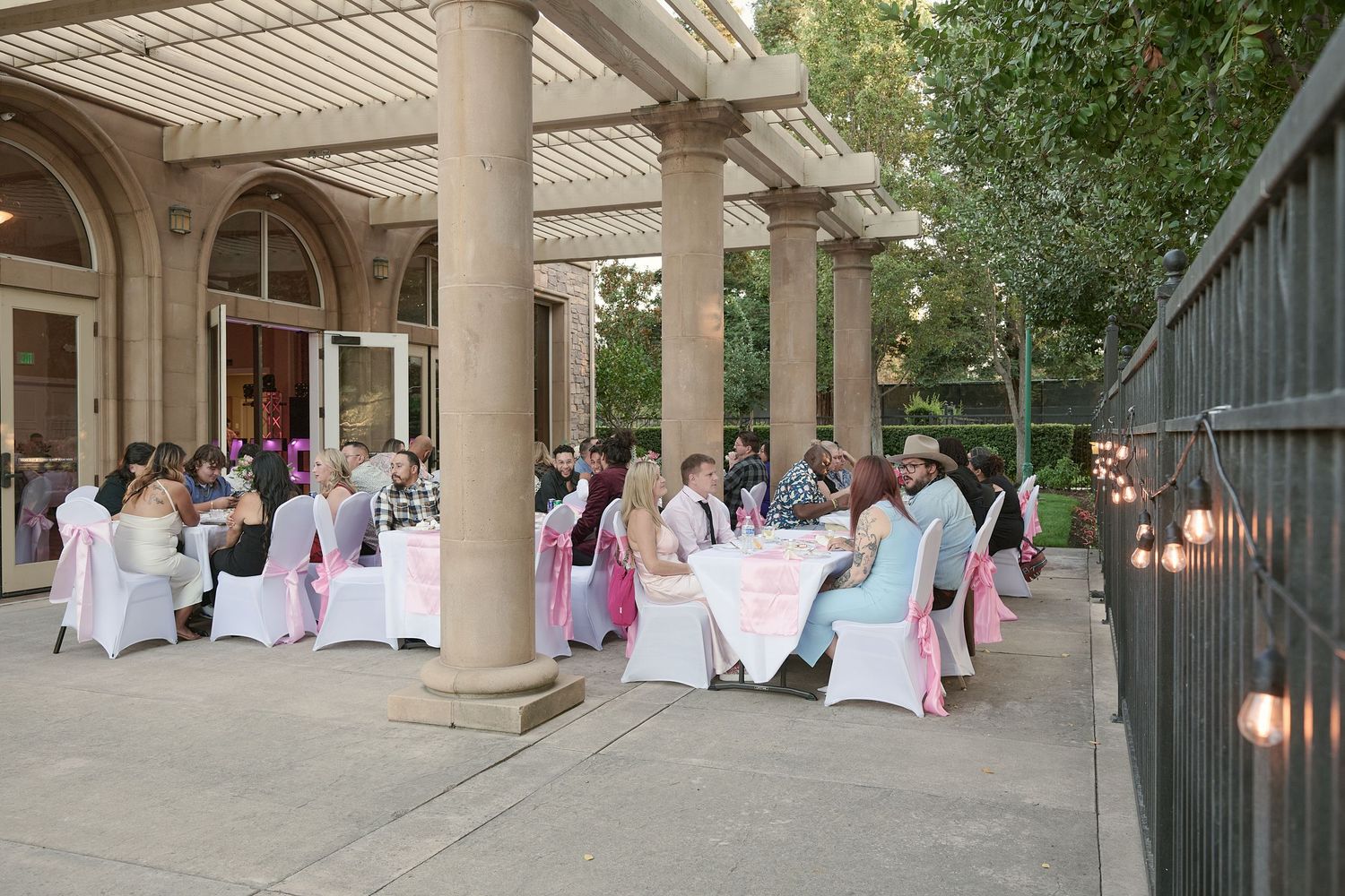 People gather under a covered walkway decorated with pink and white fabric drapes for an outdoor event.