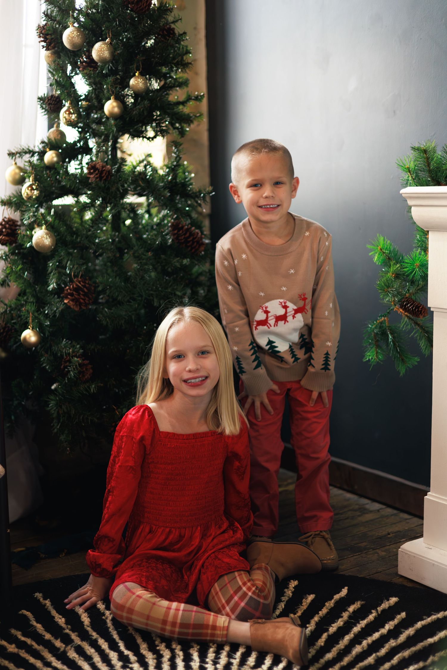 Two children in holiday outfits pose for Christmas photos in front of a decorated tree.