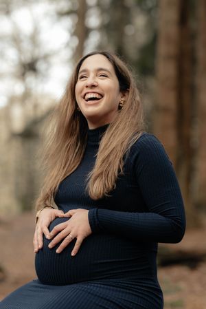 A woman in a dark sweater laughs joyfully while sitting outdoors in a wooded setting.