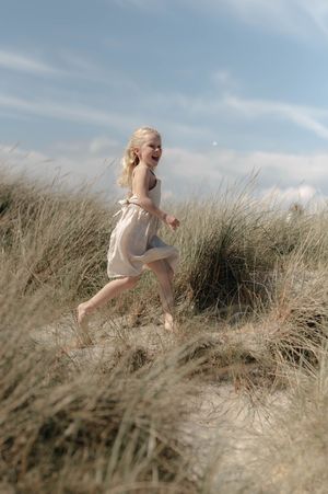 A figure in a flowing cream dress runs through tall grass against a cloudy sky in a dreamy outdoor scene.