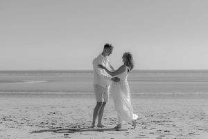 A couple in white attire dance together on an empty beach in black and white photography.