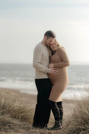 A loving couple embraces on a sandy beach with dunes and sea grass during an overcast day.