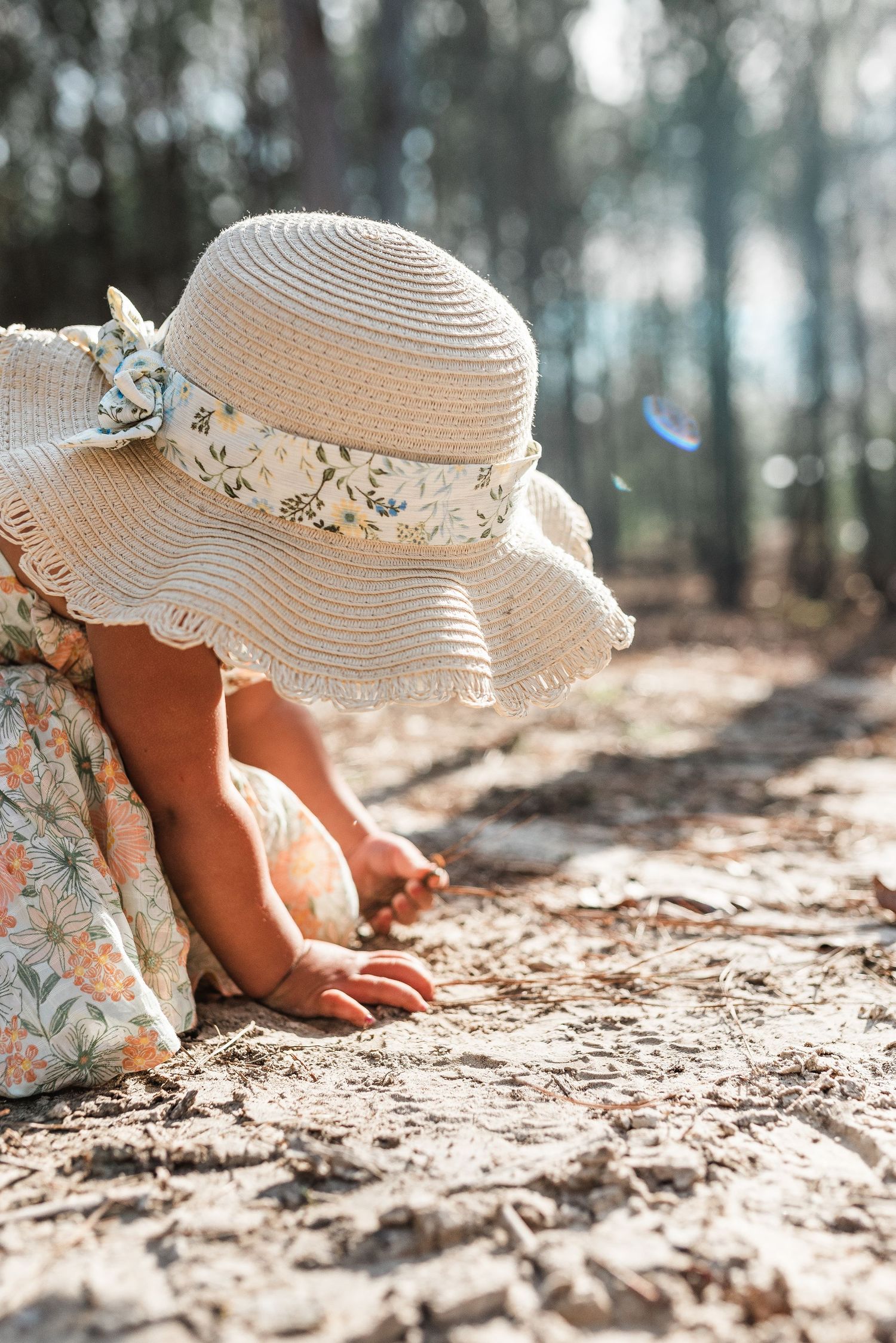 A child in a white straw sun hat with floral details plays in dirt on sandy ground in a sunlit forest setting.
