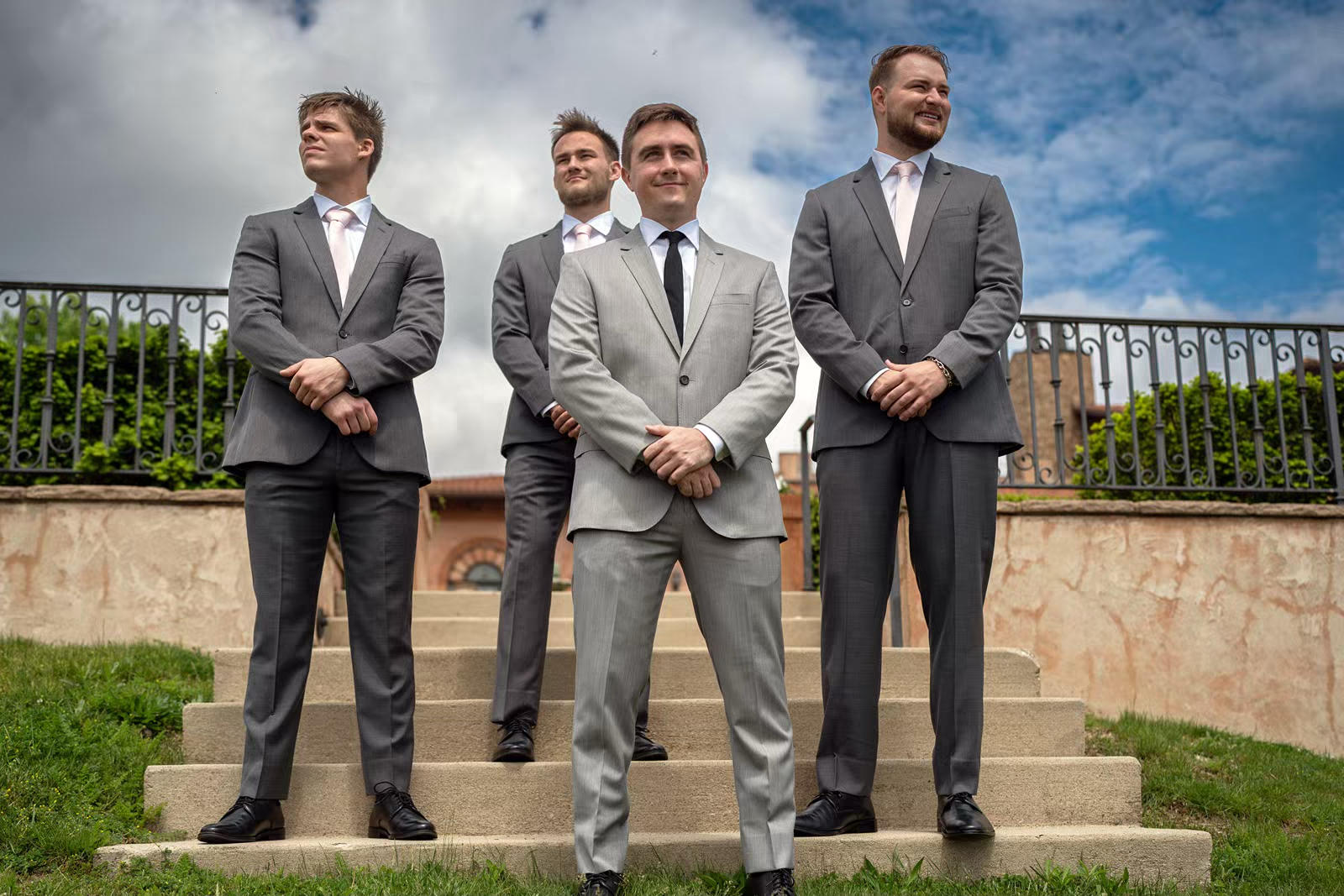 Groomsmen in matching gray suits stand on outdoor stone steps during a wedding photo session.