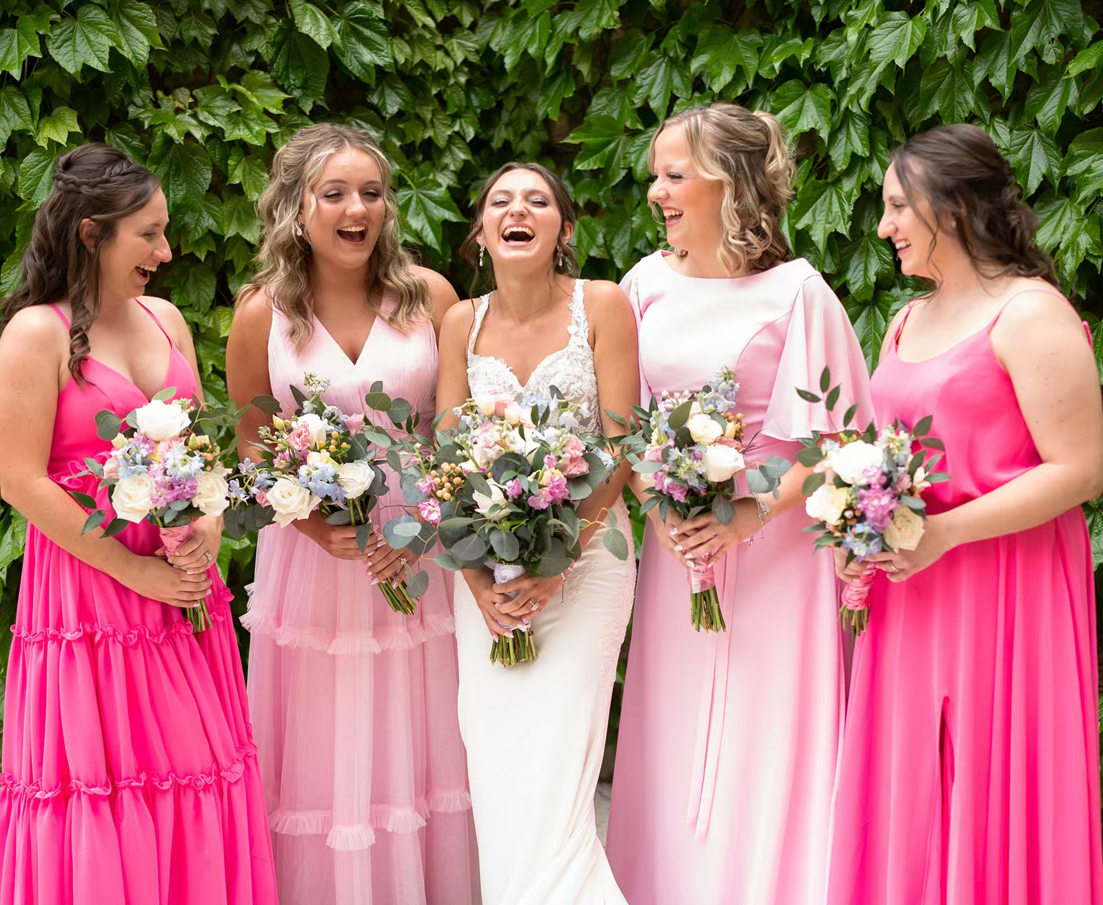 A wedding party in bright pink bridesmaid dresses and white gown stands in a line against a leafy green backdrop.