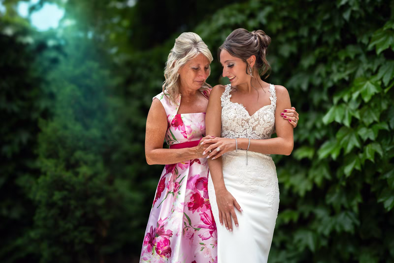 Two people in formal attire embrace outdoors against a lush green foliage backdrop.
