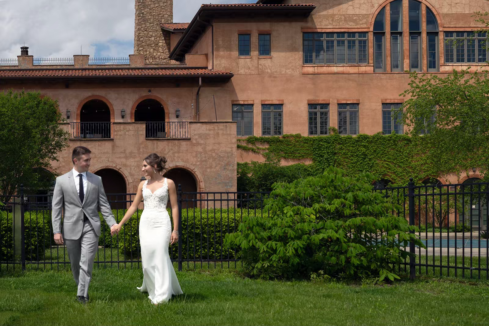 A couple in wedding attire walks together across a lush green lawn in front of a historic brick building.
