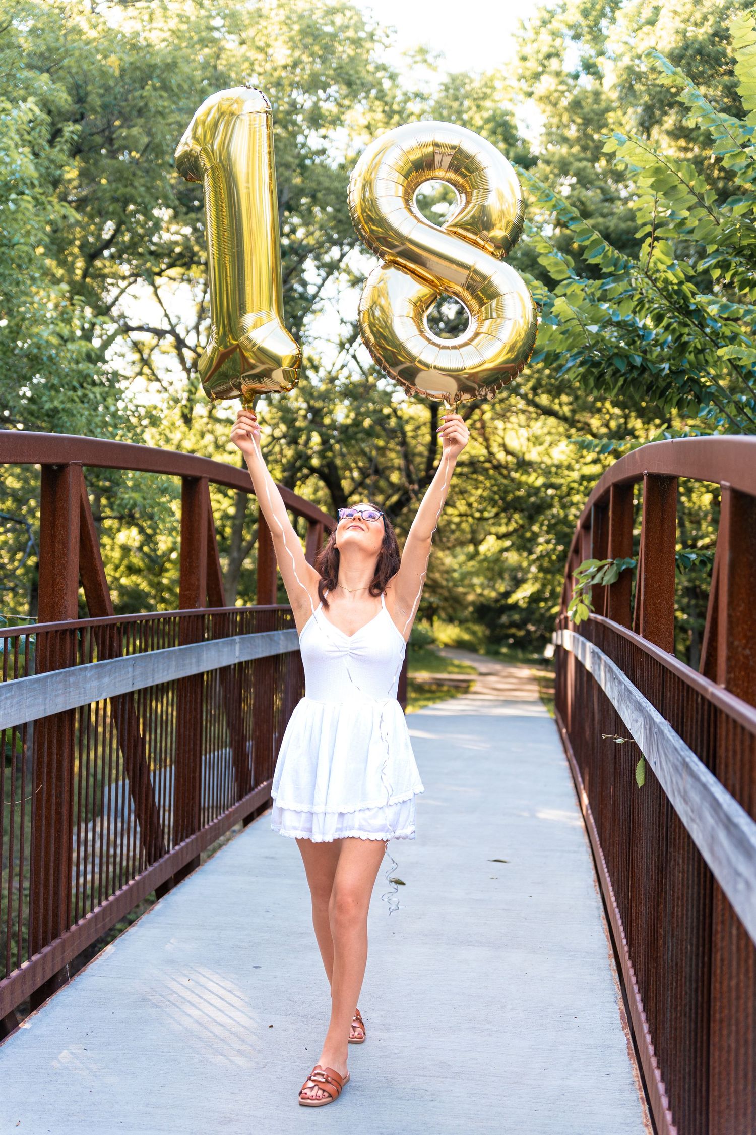 Graduate in white dress holds gold number balloons while celebrating on wooden bridge.