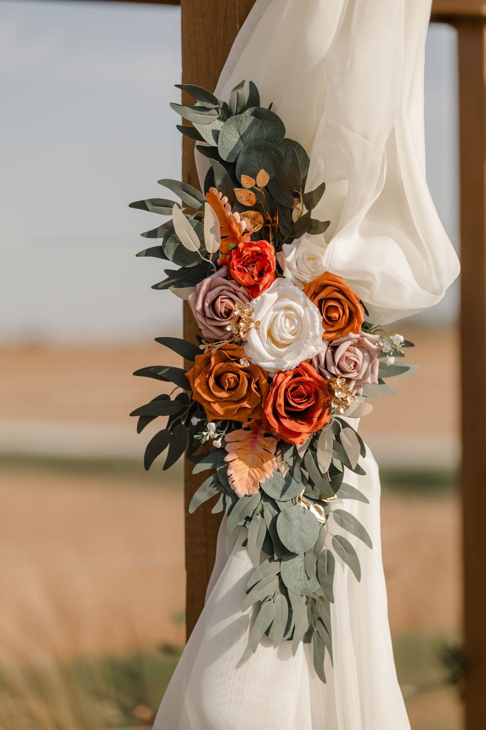White fabric draping adorned with a romantic floral arrangement featuring peach, orange and rust-colored roses with eucalyptus leaves.