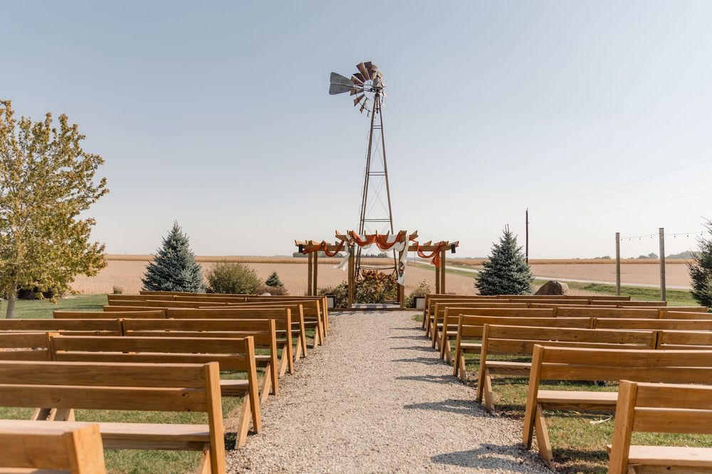 Wooden pews line a pathway leading to a rustic windmill under a golden sunset sky.
