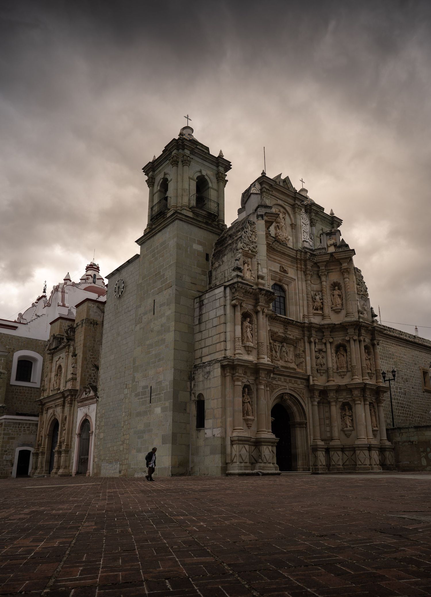 Ancient stone cathedral facade with ornate Baroque architecture under dramatic cloudy sky in Oaxaca, Mexico.