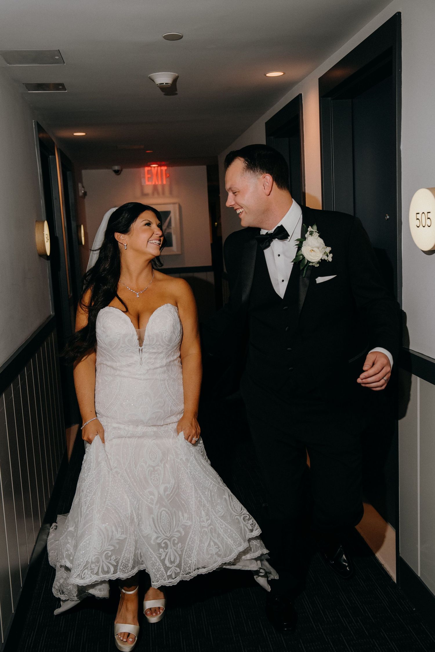 A couple in formal wedding attire shares a playful dance moment in a dimly lit hallway.