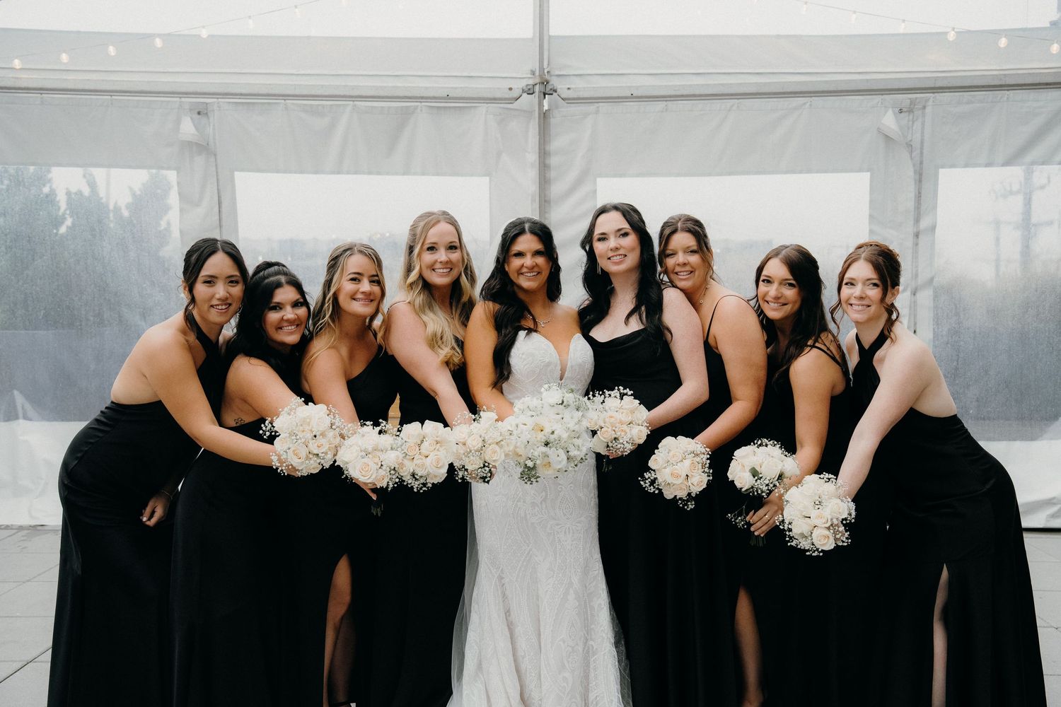 A wedding party wearing black dresses and holding white bouquets poses together for a formal group photo.
