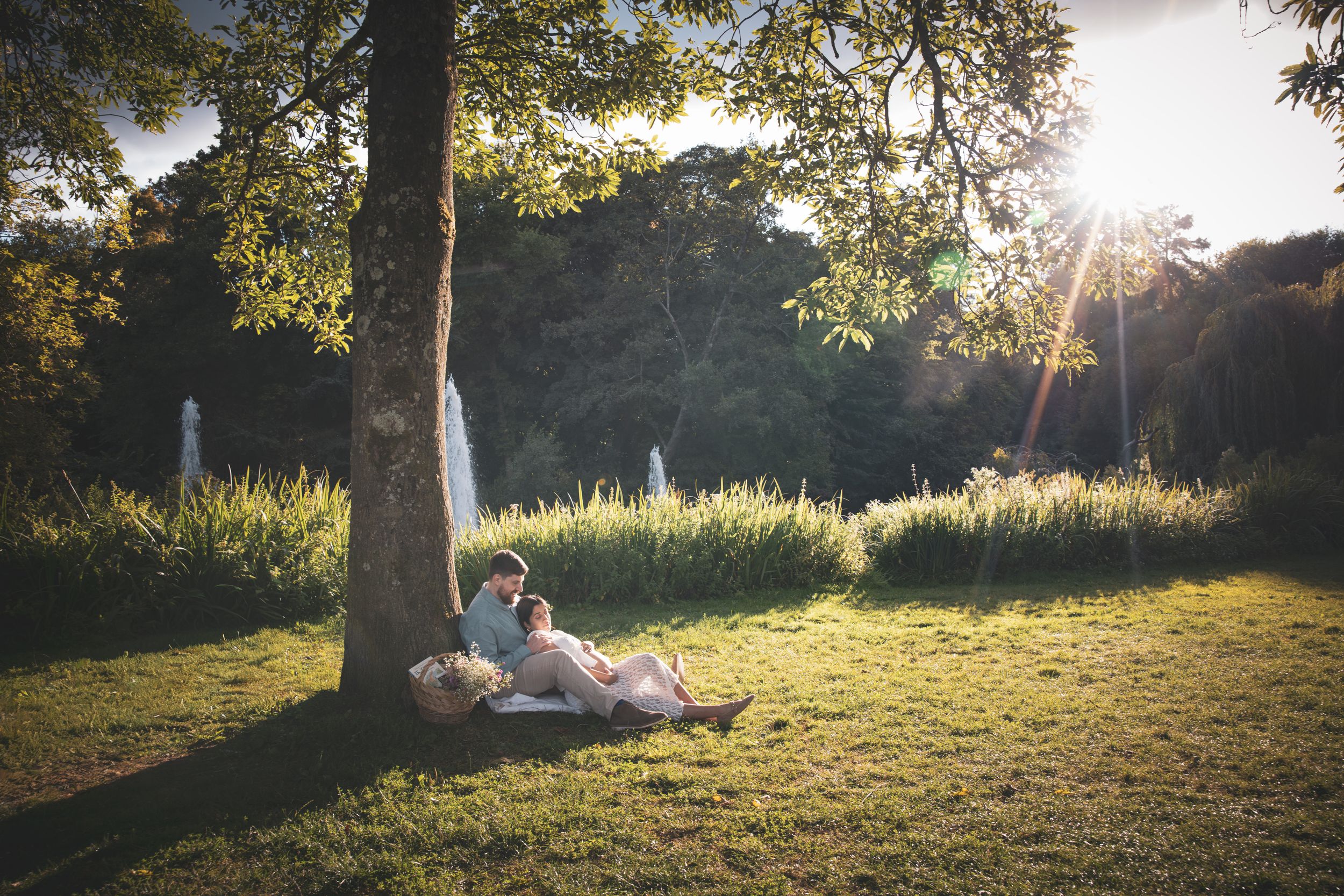 A couple lounging against a tree, sunlight streams through trees in a tranquil meadow during golden hour creating a warm atmospheric scene.