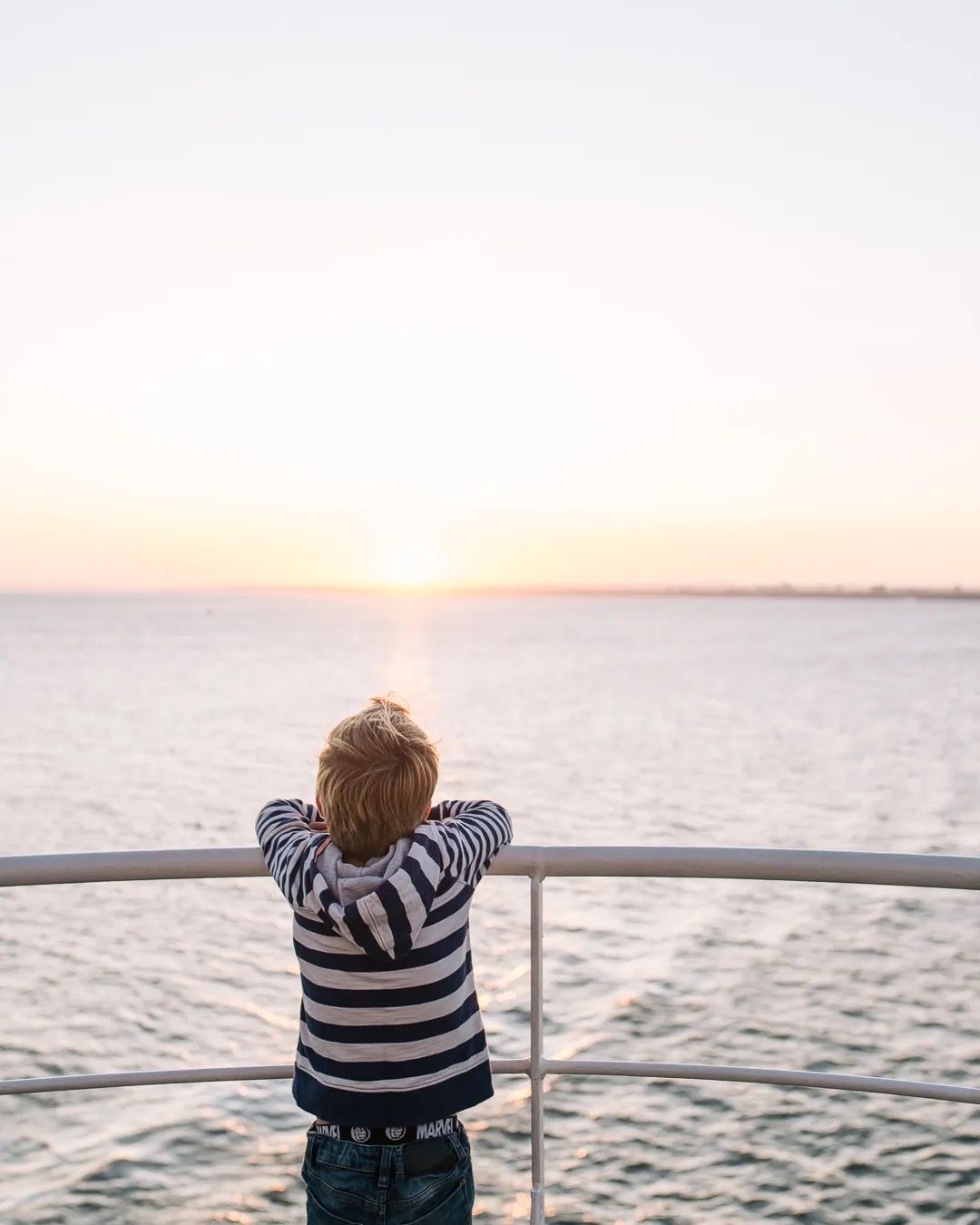 Child standing at a ferry railing looking out at the sea during sunset.