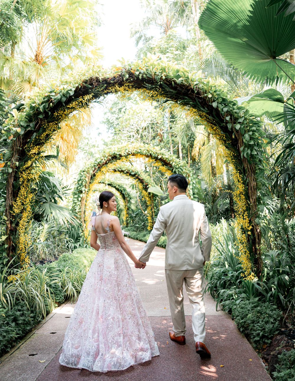 A couple walks under a lush garden archway decorated with yellow flowers and greenery in a romantic outdoor setting.