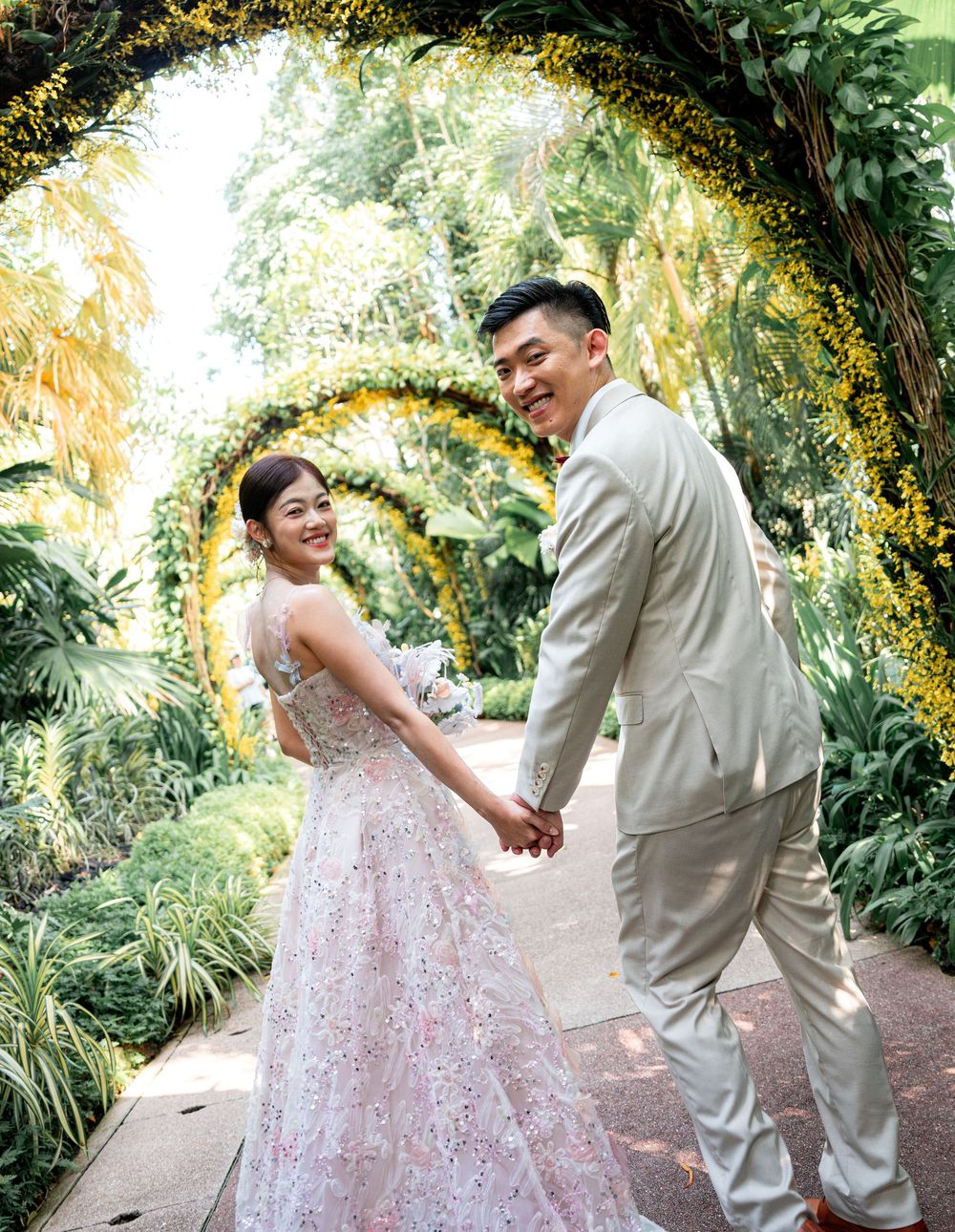 Couple in wedding attire dance together under lush garden archway with palm trees and greenery in background.