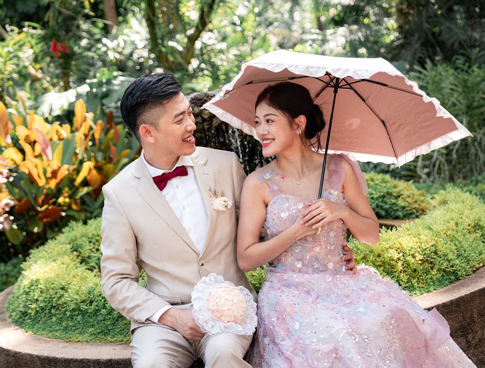 A couple in wedding attire shares a romantic moment under a pink umbrella in a lush garden setting.
