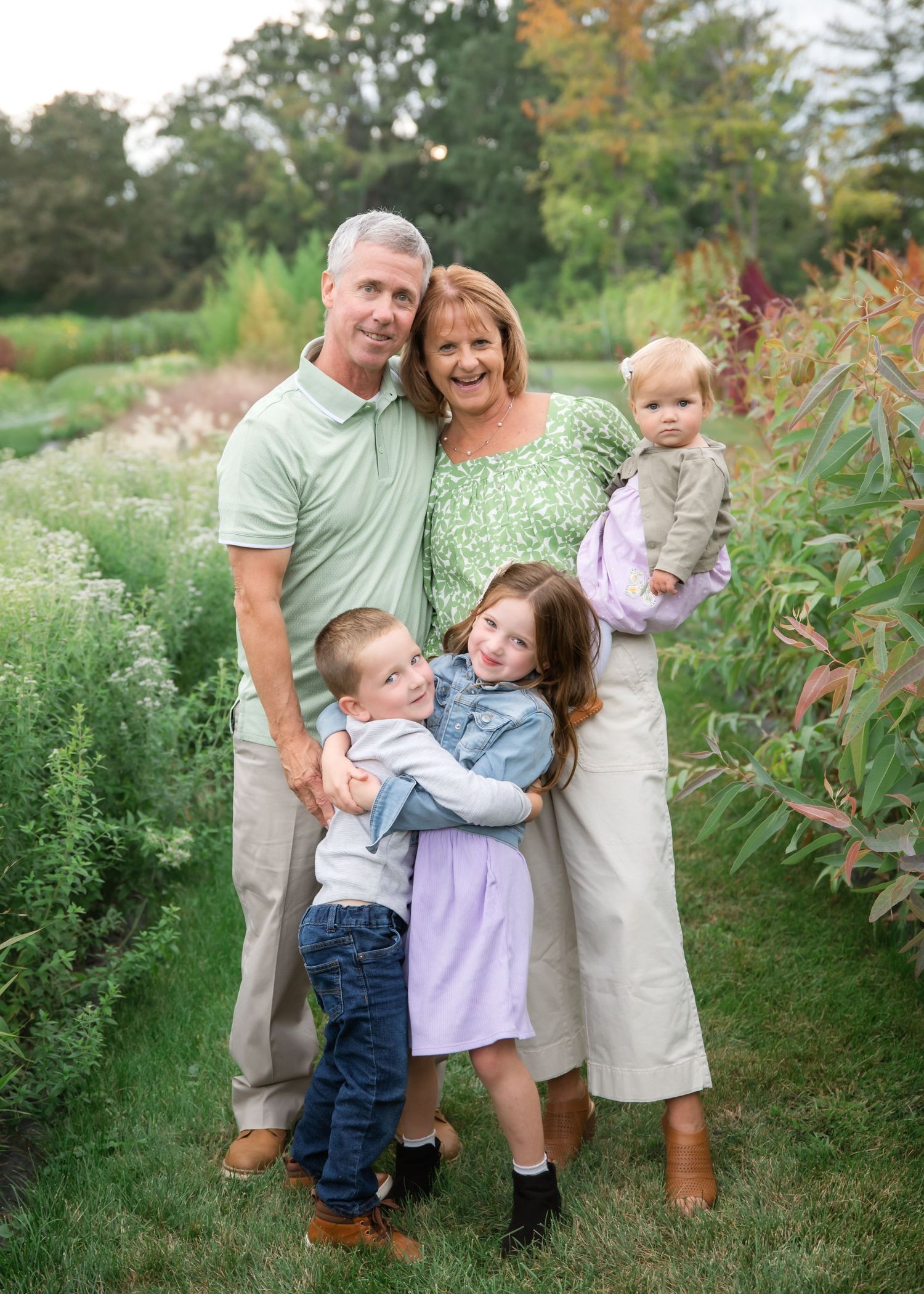A family of four poses together outdoors in coordinated casual outfits amidst lush greenery during a summer photo session.