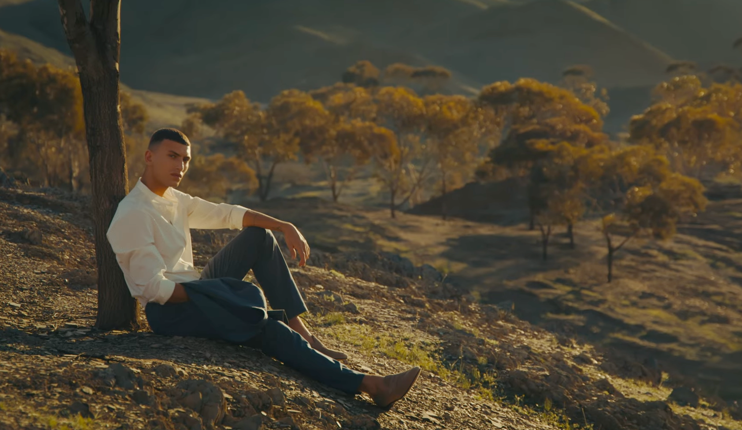 A couple sits together on rocky terrain during golden hour as sunlight casts warm shadows across the landscape.