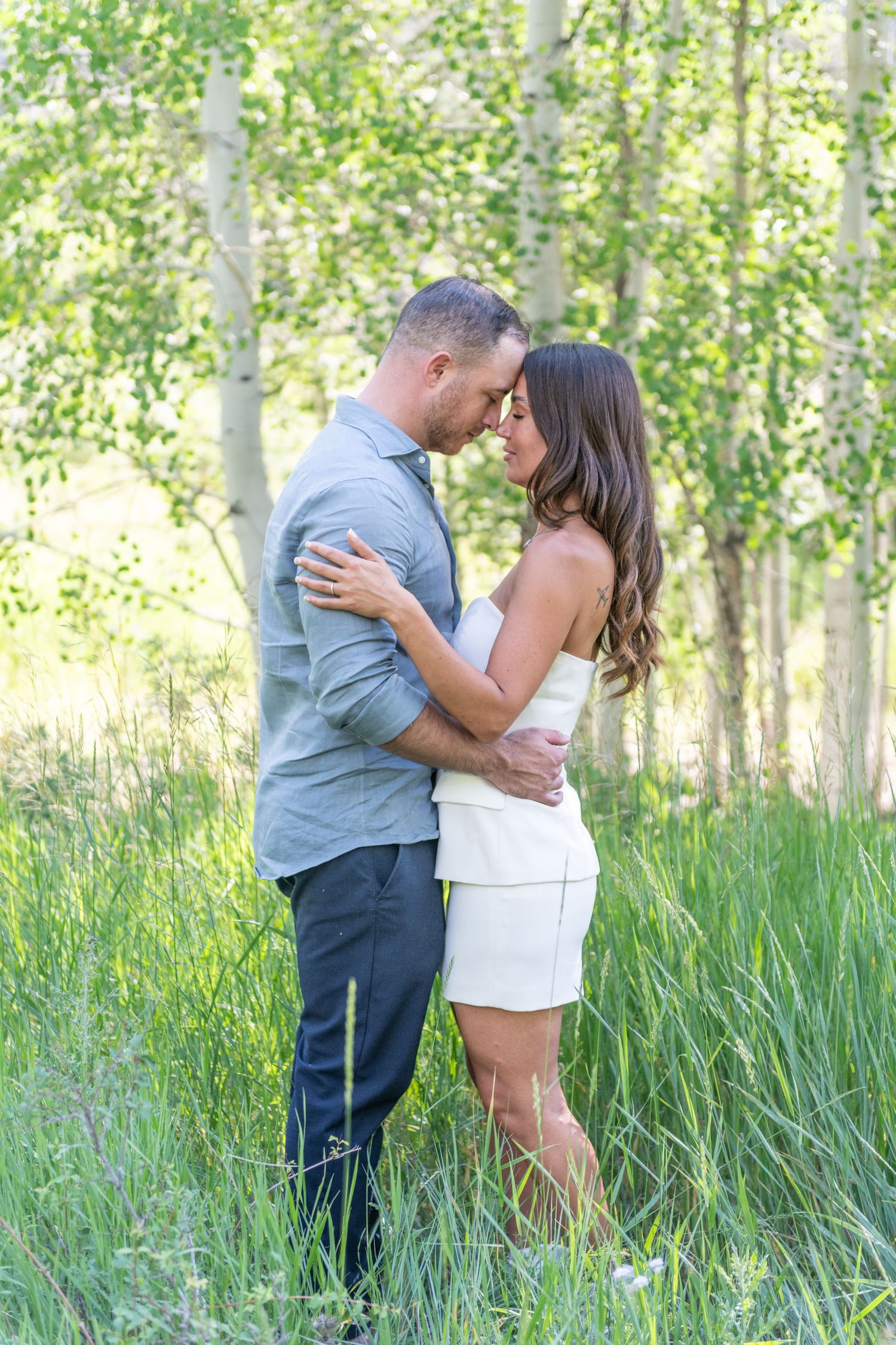 A bride and groom are kissing in a mountain meadow. The meadow is filled green aspen and white  flowers with mountains in the background.