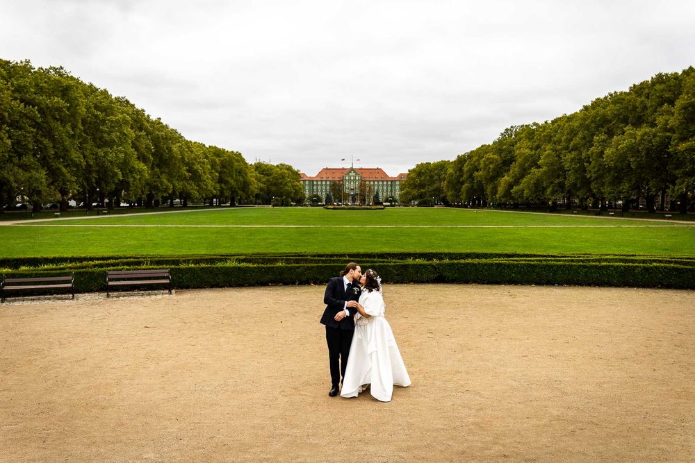 A couple in wedding attire stands on a sandy path with a grand mansion and green lawn in the background.