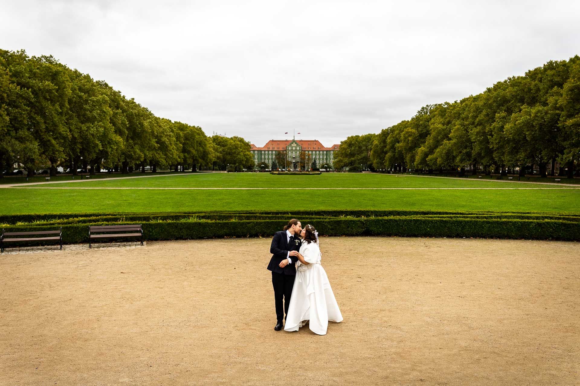 A couple in wedding attire stands on a sandy path with a grand mansion and green lawn in the background.