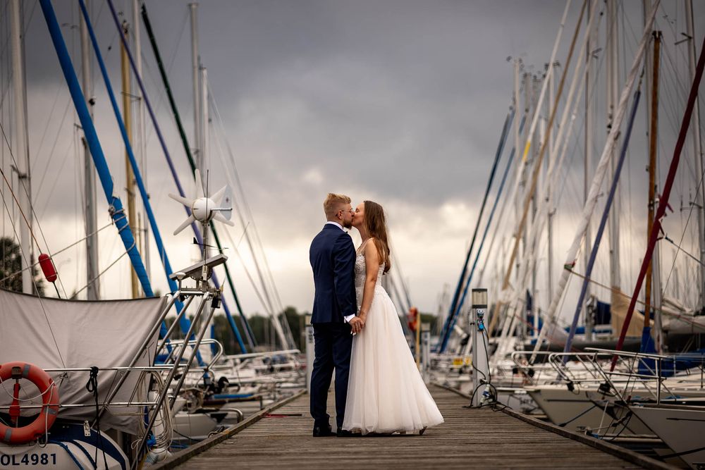 A couple shares a romantic moment on a marina dock surrounded by sailboat masts against a dramatic stormy sky.