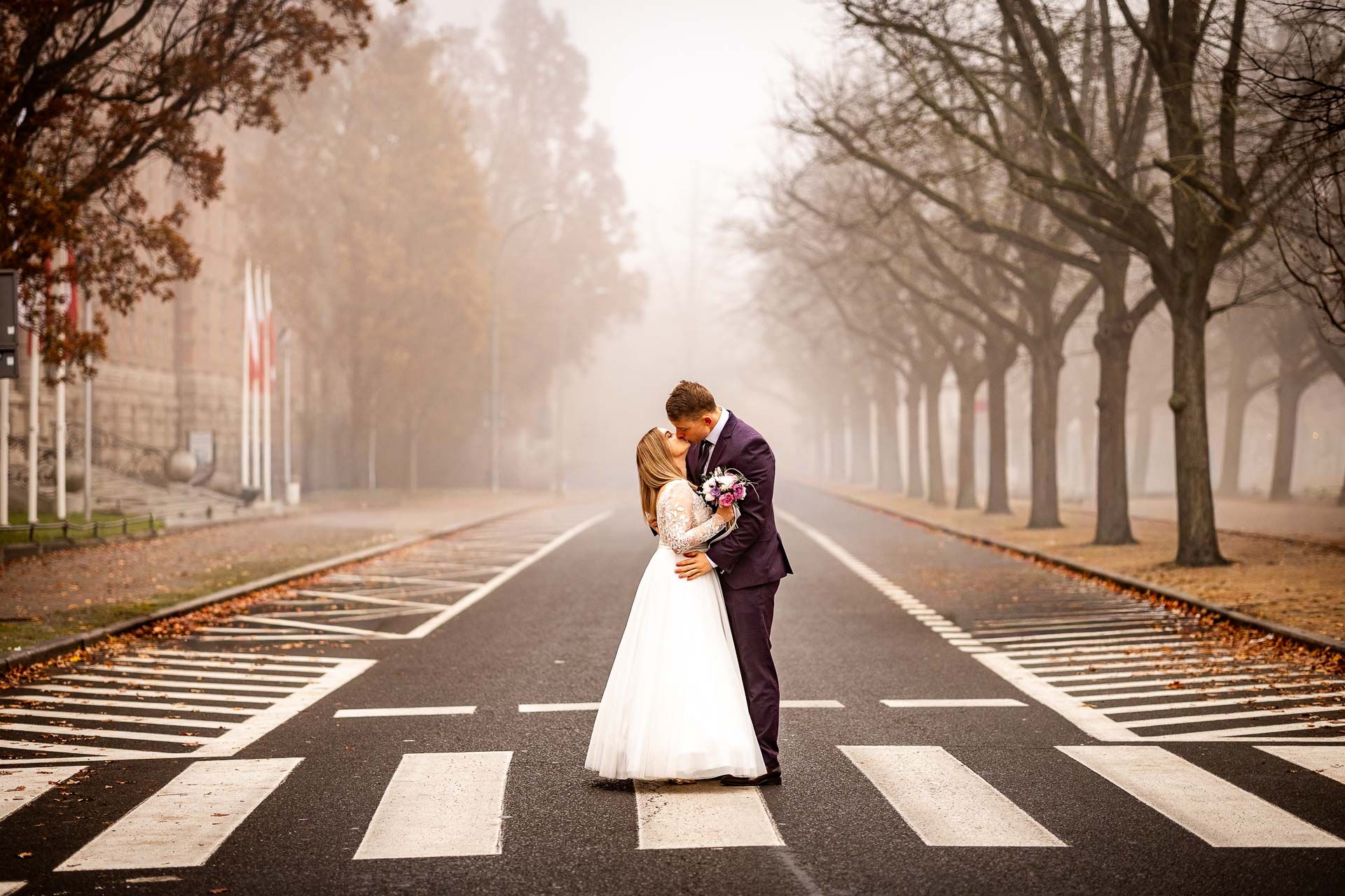 A couple in wedding attire embraces on a foggy tree-lined street with crosswalk stripes in the foreground.