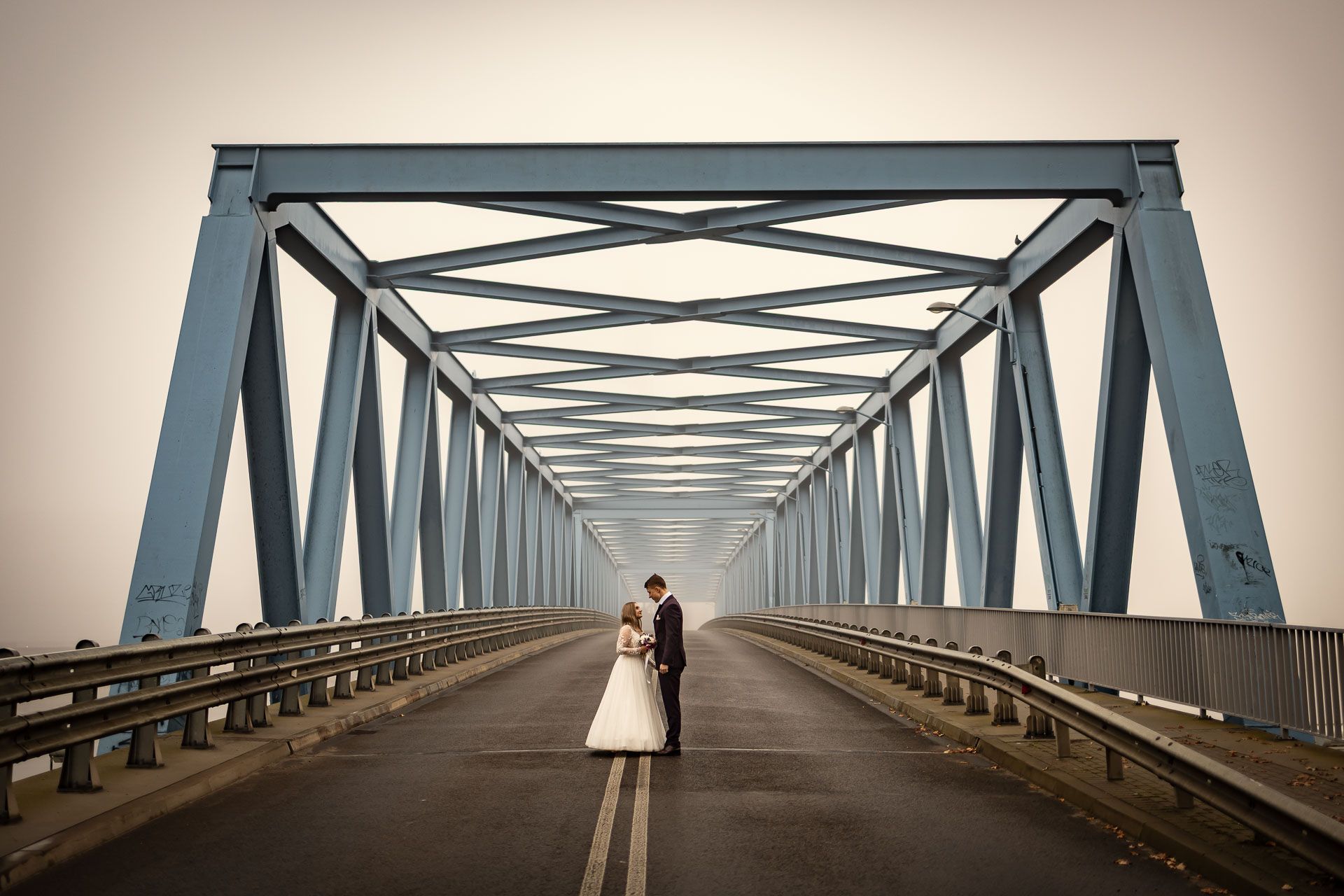 A couple stands in the center of a steel bridge tunnel with dramatic architectural lines leading to a vanishing point.