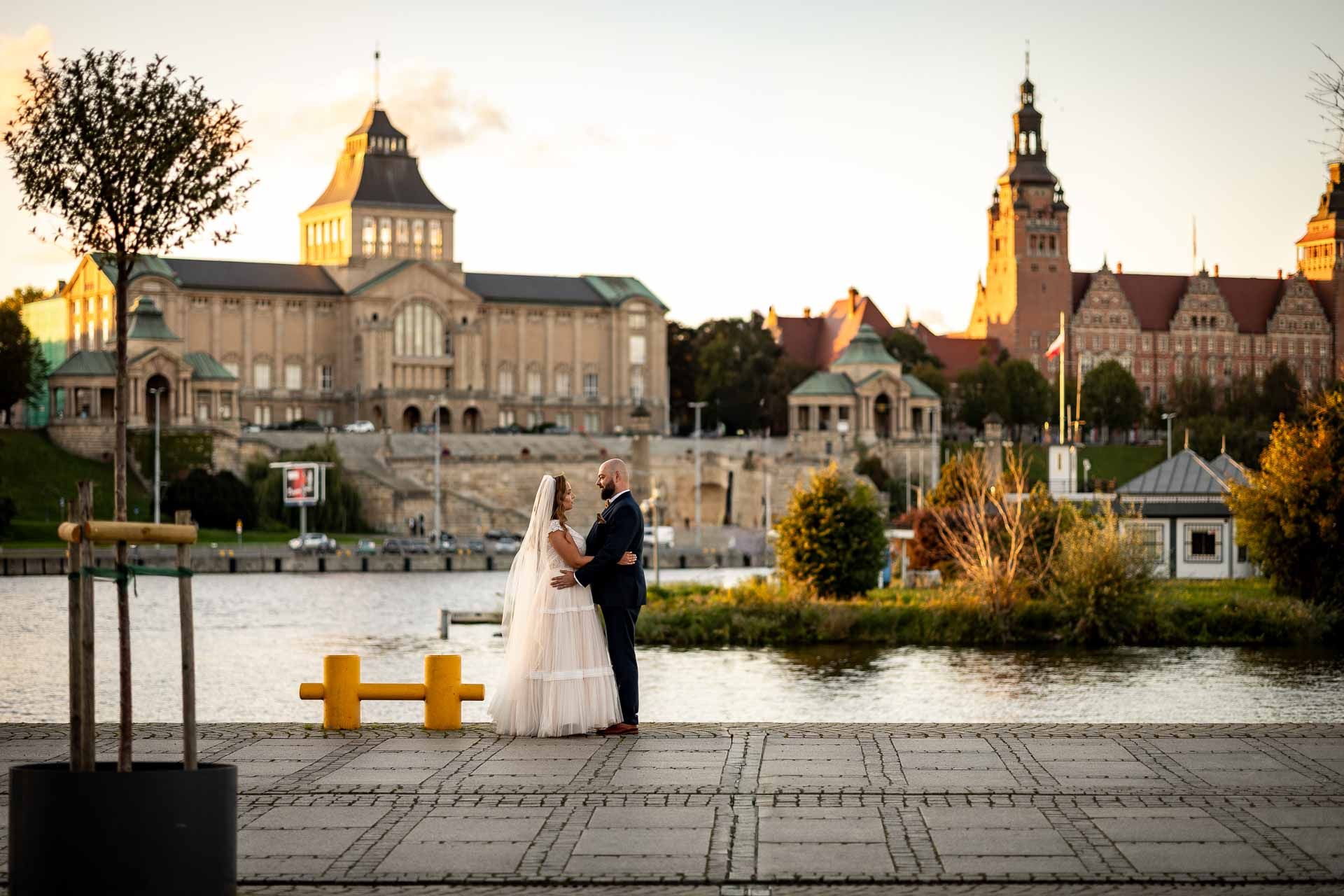 Couple in wedding attire poses at sunset in front of historic European architecture and waterfront buildings.