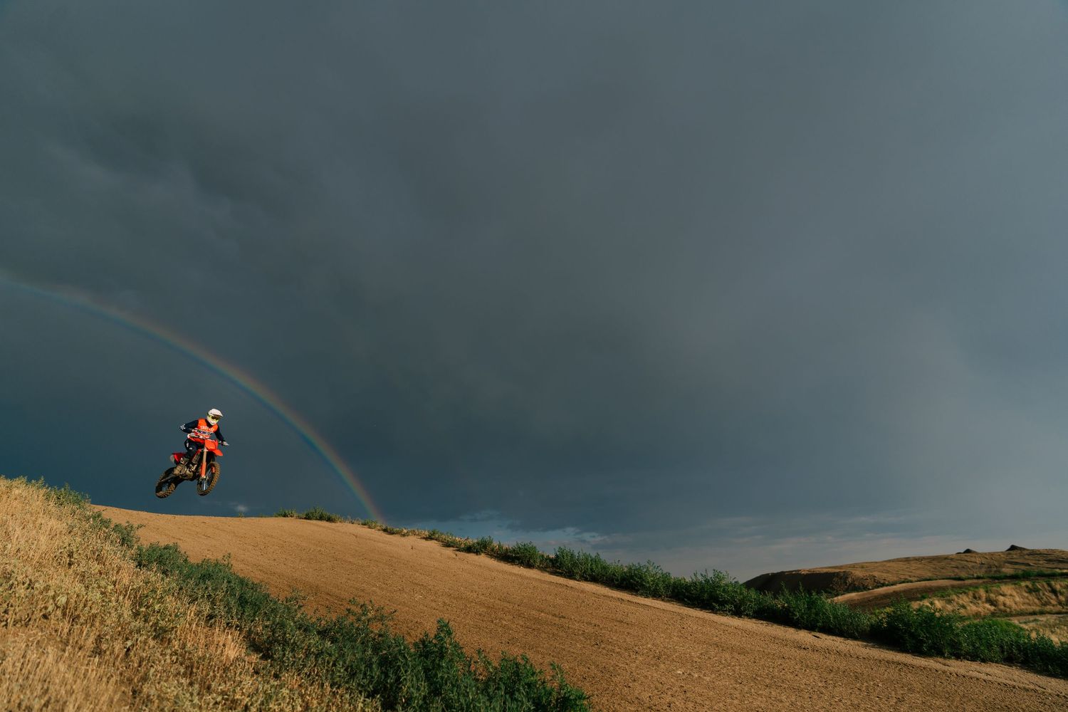 Motorcyclist rides along scenic hilltop trail against dramatic stormy sky during sunset.