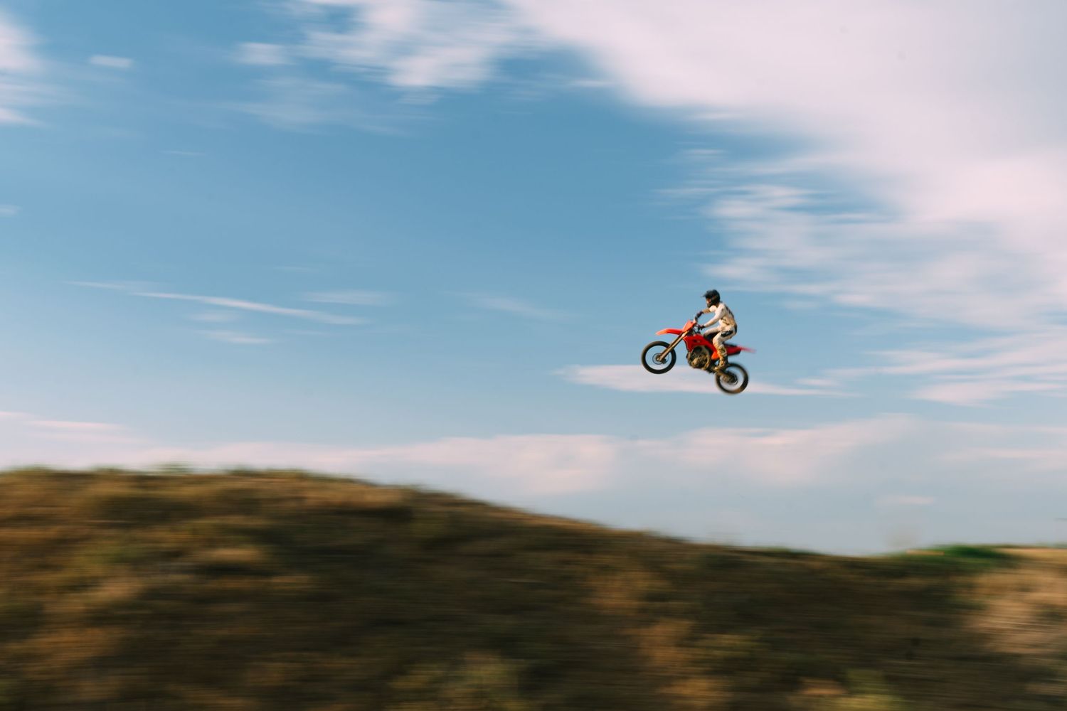 A motocross rider soars through the air on a dirt bike against a blue sky with rolling hills in the background.