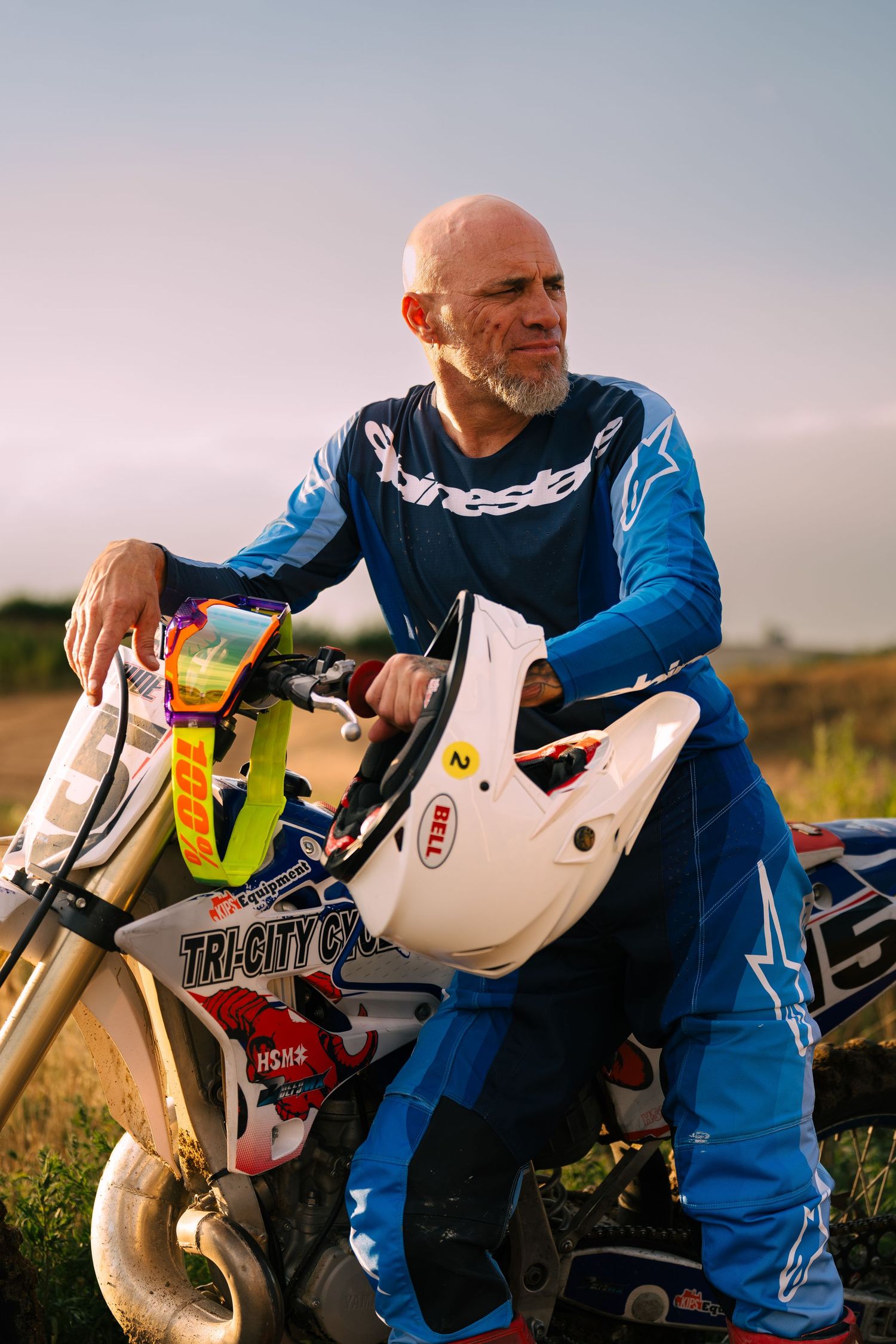 A motocross rider in a blue jersey takes a break beside their bike in a grassy field at sunset.
