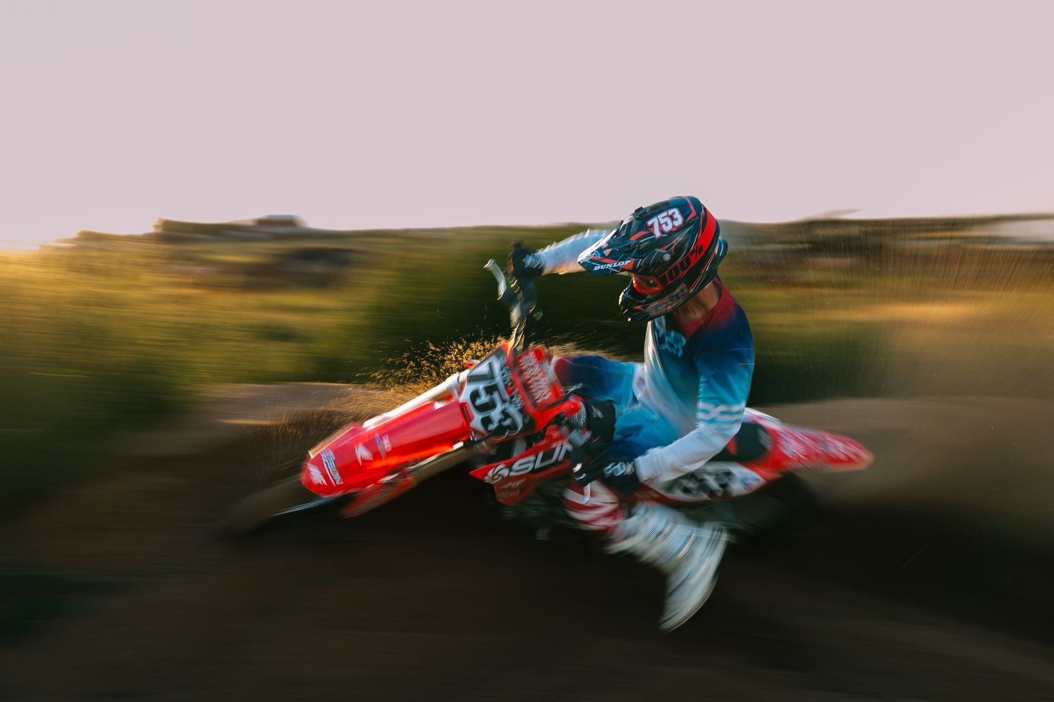 Motocross rider performing a dramatic sideways whip on a red dirt bike in motion against blurred landscape.