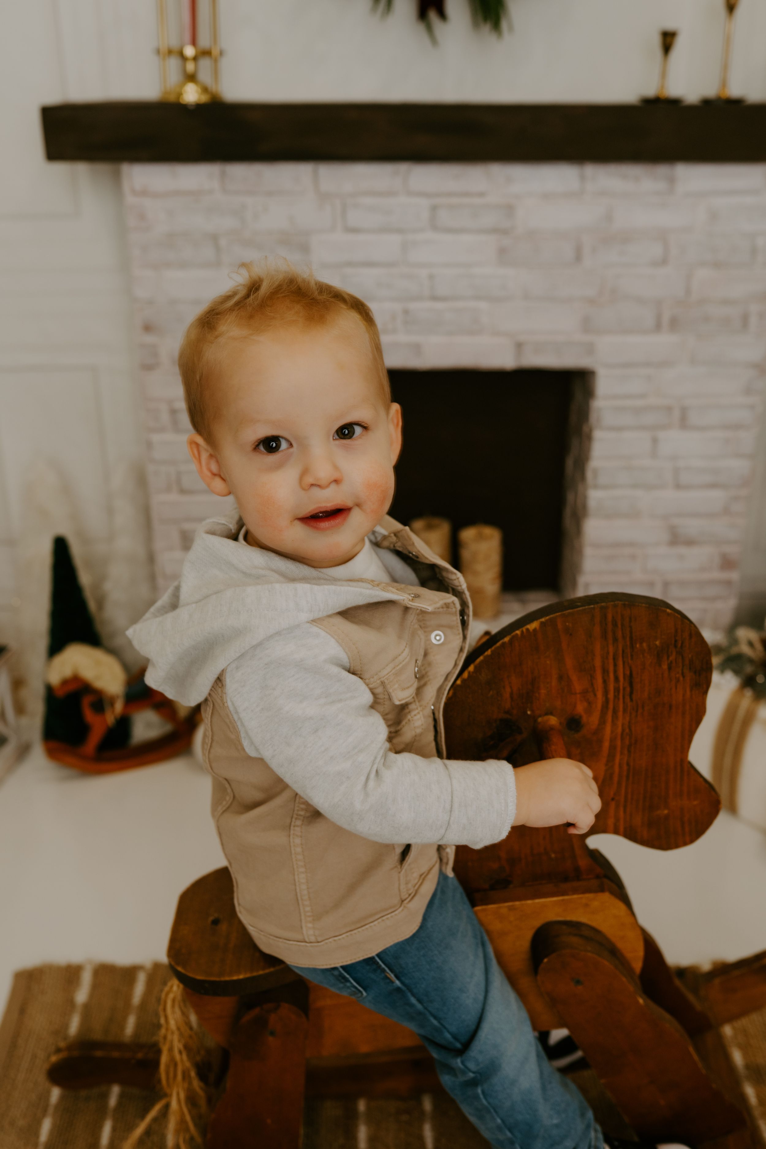 Baby in tan vest and jeans plays on wooden rocking horse near white brick fireplace.