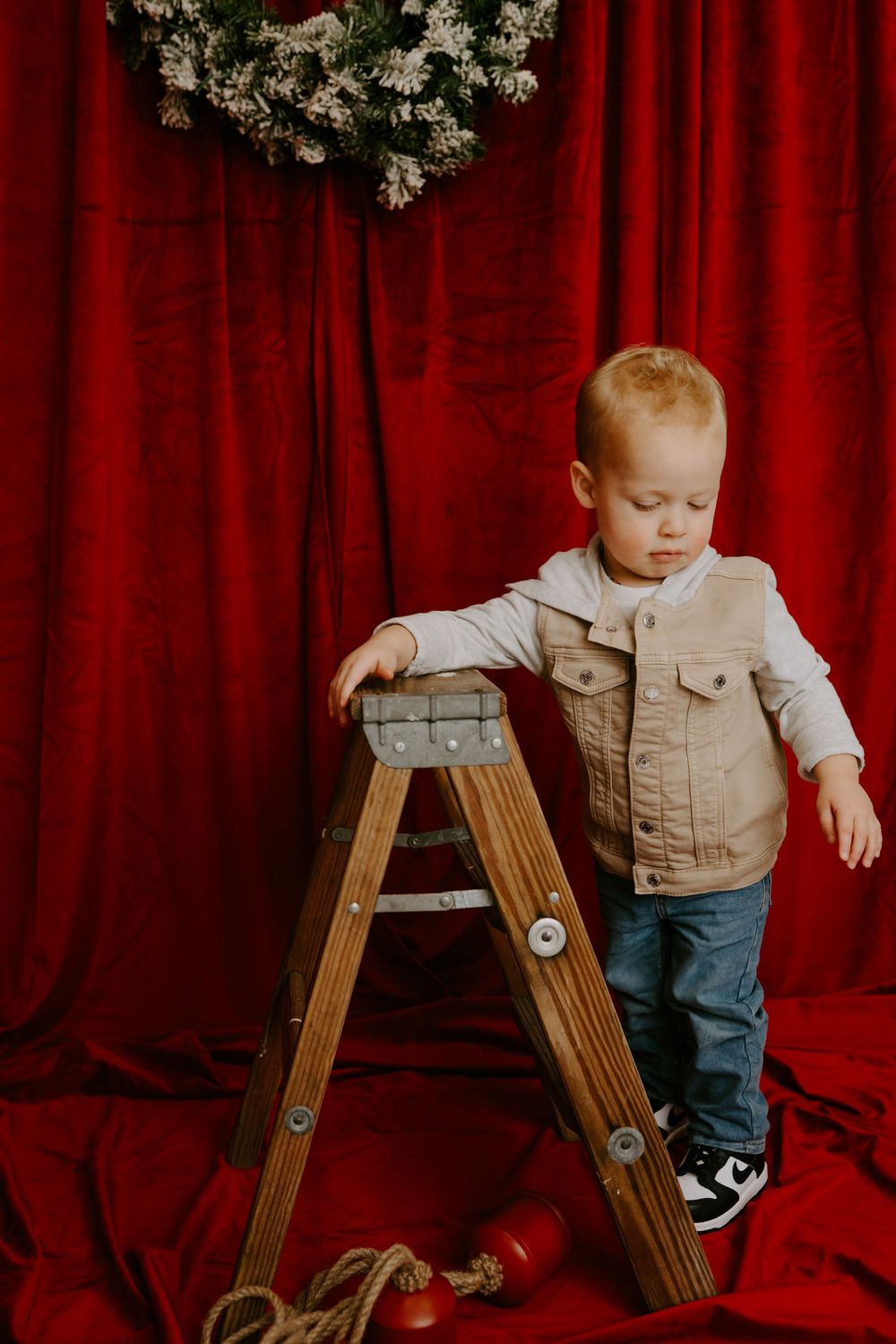 Young child in tan vest and jeans explores wooden ladder against red curtain backdrop.