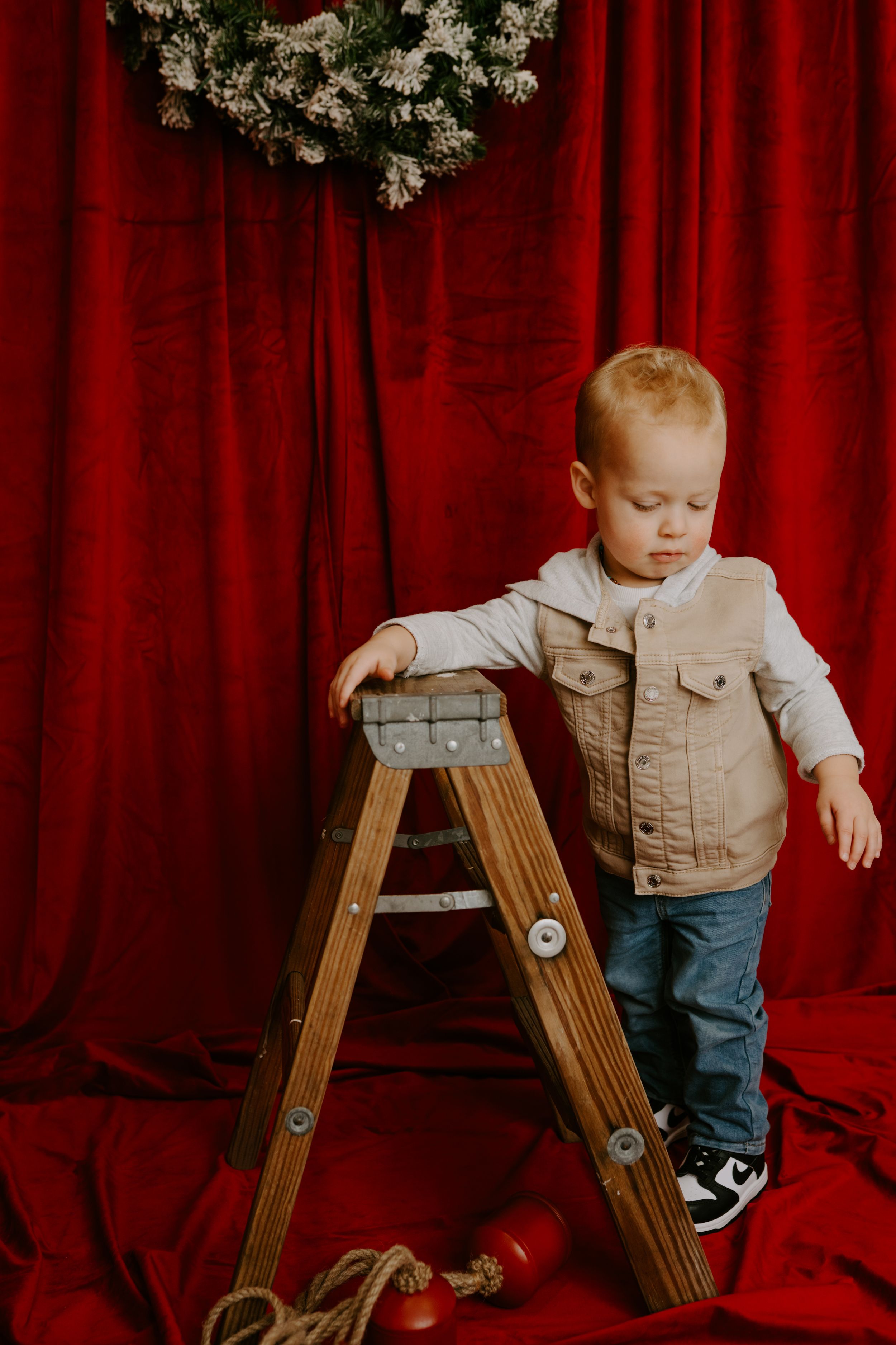 Young child in tan vest and jeans explores wooden ladder against red curtain backdrop.