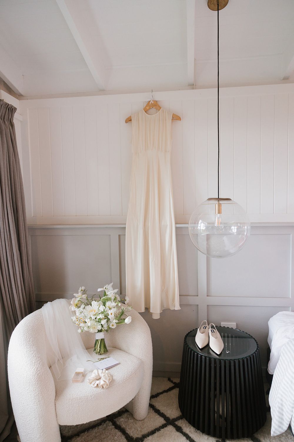 A cream-colored dress hangs above a white curved chair with a bouquet and black side table in a modern bedroom setting.
